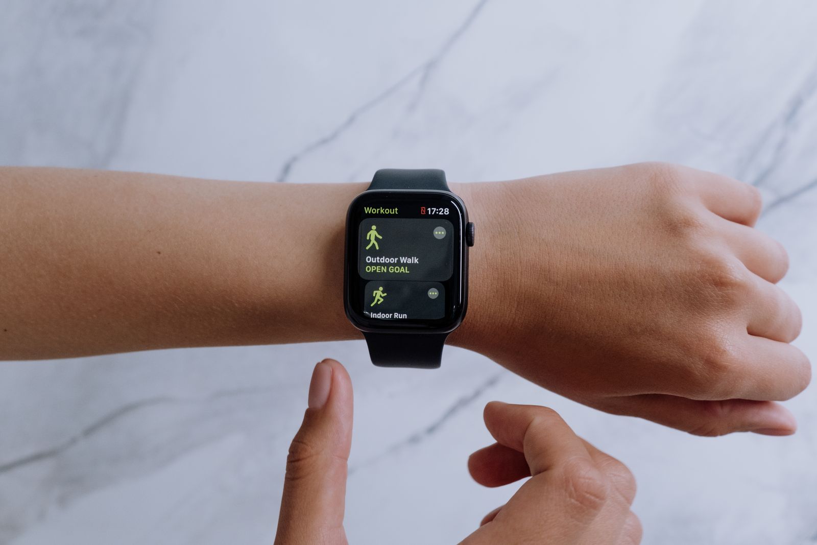 Smartwatch de pulseira preta no pulso, com a tela de treinos aberta, sobre fundo de mármore claro